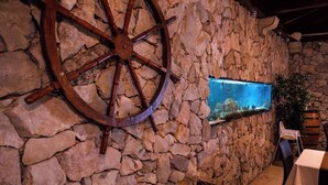 A stone wall with a large wooden wheel, a fish tank, and a rustic setting with tables and chairs. - Holiday apartment Sara with sea views and pool (Klimno)