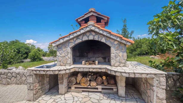 A stone-built outdoor fireplace with a tiled roof, surrounded by a stone wall and a paved area.