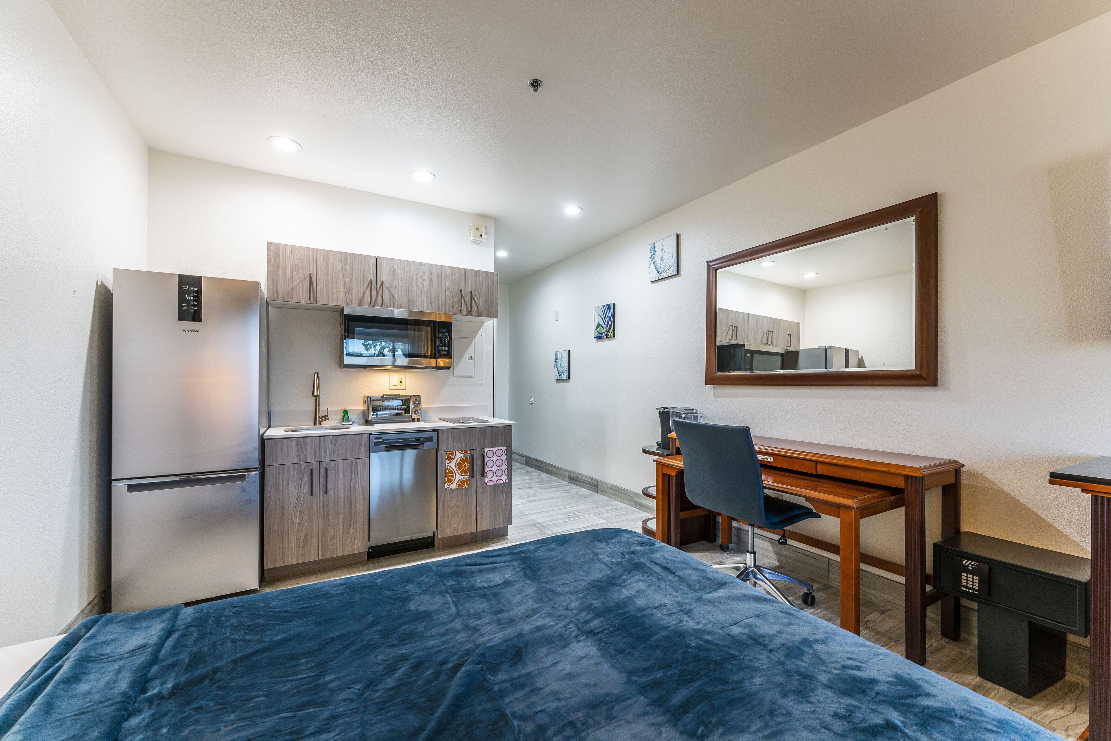 A compact room with a bed, a desk, a chair, a small kitchenette, and a mirror.