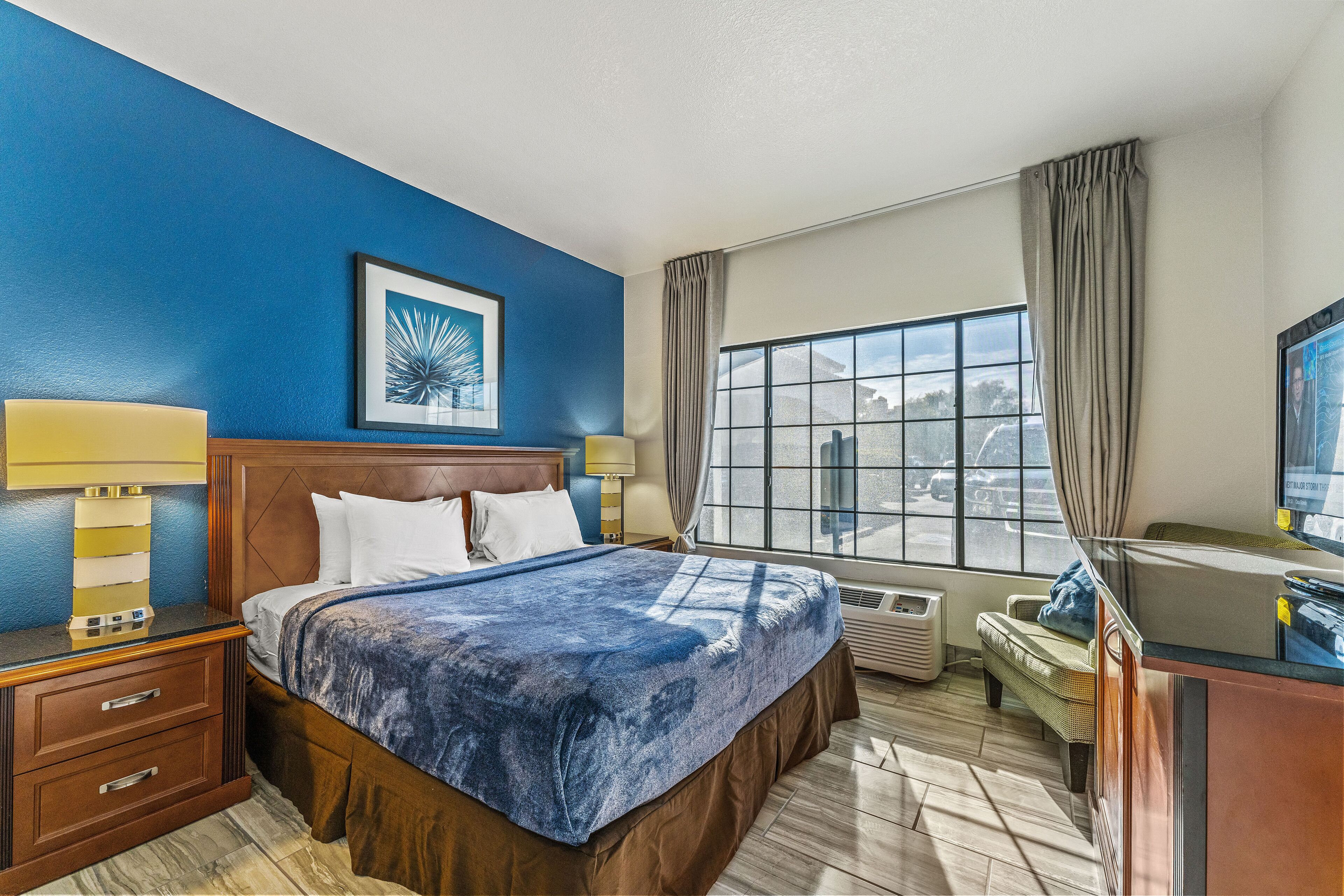 Een hotelkamer met een groot bed, een bureau met een computer, een raam met gordijnen en een lijst met een afbeelding aan de muur.