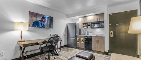 A compact hotel room with a kitchenette, a TV mounted on the wall, a desk with a chair, and a sofa.