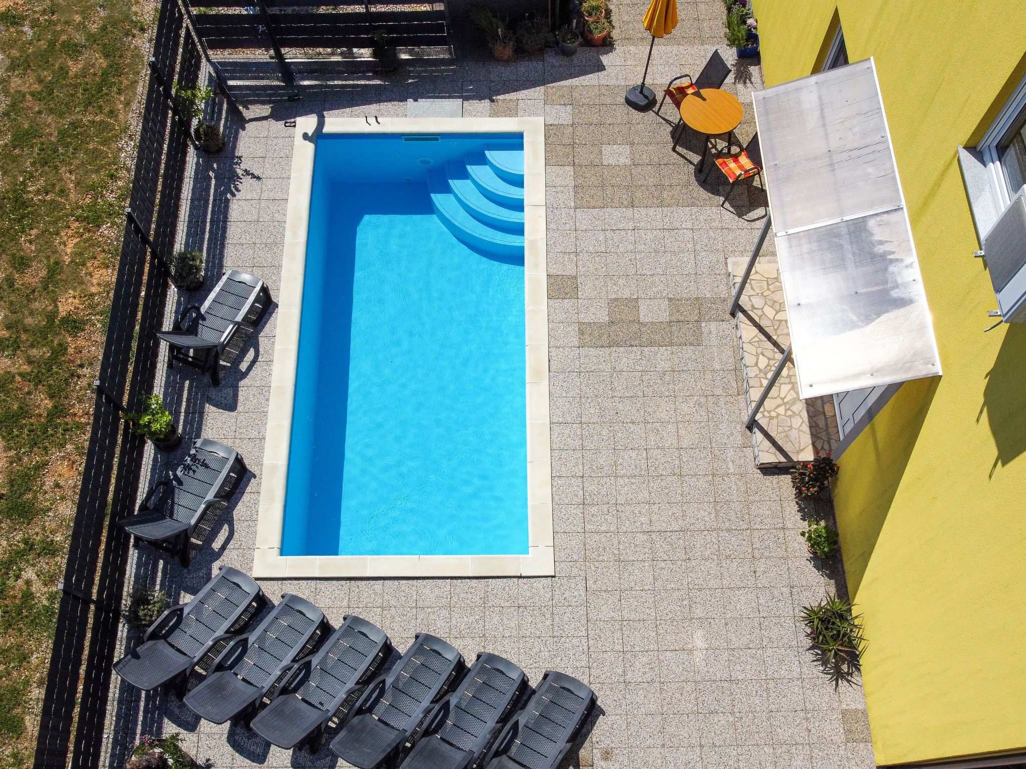 Une piscine rectangulaire entourée d’une terrasse carrelée. On y trouve plusieurs chaises longues et une petite table avec un parasol jaune.