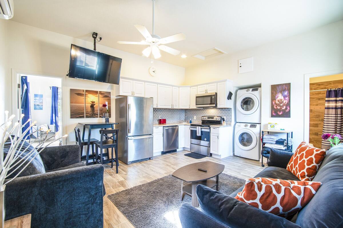 A modern kitchen with stainless steel appliances, a dining area with a table and chairs, and a living room with a sofa and a ceiling fan.