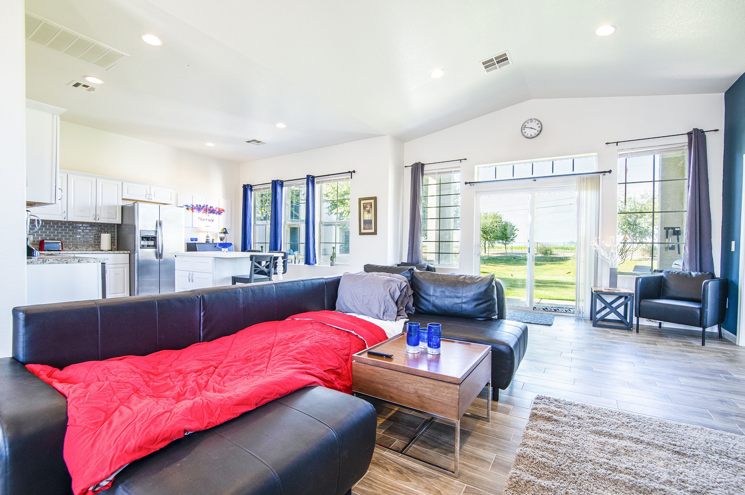 A spacious living area with a black leather sofa, a red blanket, and a coffee table. There is a kitchen in the background with white cabinets and a refrigerator.