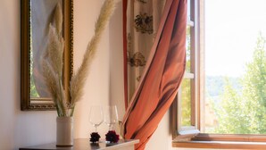 Ein Zimmer mit Kommode, einer Vase mit Pampasgras, zwei Weingläsern und einem Fenster mit Vorhängen.