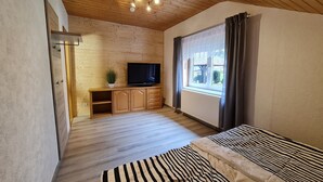 A bedroom with a wooden ceiling, a TV on a wooden cabinet, a window with curtains, and a striped bed. - Vacation in nature - view of the Elbe Sandstone Mountains in Saxon Switzerland (Königstein)