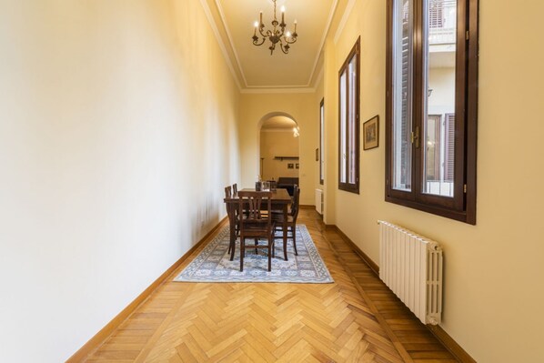 A hallway with a dining table, chairs, a patterned rug, a radiator, and a chandelier.