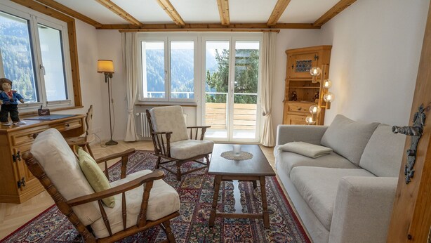Una sala de estar acogedora con muebles de madera, un tapete estampado y vistas a las montañas a través de amplias ventanas.