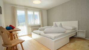 Un dormitorio moderno con cama, una silla de madera, una mesita pequeña y vistas al exterior a través de amplios ventanales.