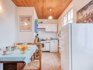 Une cuisine de taille réduite avec un plafond en bois, des placards blancs, une table à manger recouverte d’une nappe bleue et un réfrigérateur.