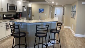 Una cocina moderna con una isla central, taburetes de bar, electrodomésticos de acero inoxidable y vistas al salón.