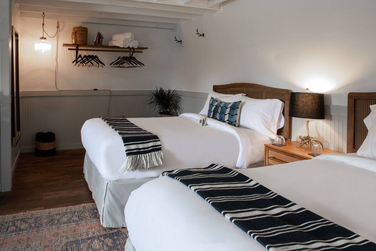 A hotel room with two beds, a wooden headboard, a bedside table with a lamp, a hanging light fixture, a plant, and a shelf with towels and hangers.