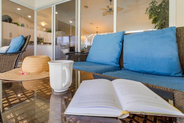 A modern living area with a glass table, a wicker sofa with blue cushions, a straw hat, and a white mug.