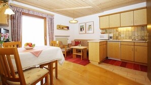Een gezellige kamer met een eettafel, stoelen, een sofa, een kleine keuken met kastjes en een spoelbak, en een raam met gordijnen.