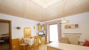 Een gezellige kamer met houten balkenplafond, een zithoek met stoelen en een tafel, een keuken met aanrecht en een raam met gordijnen.