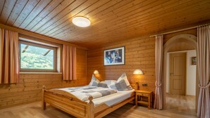 A wooden-paneled bedroom with a bed, a nightstand, a wall-mounted lamp, a window with curtains, and a door with an archway. - Charming holiday home near the ski resort (Kaltenbach)