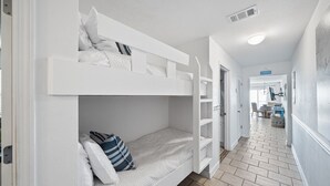 Un lit superposé avec une literie blanche et des oreillers à rayures, situé dans un couloir dont le sol est carrelé et qui mène à une salle à manger.