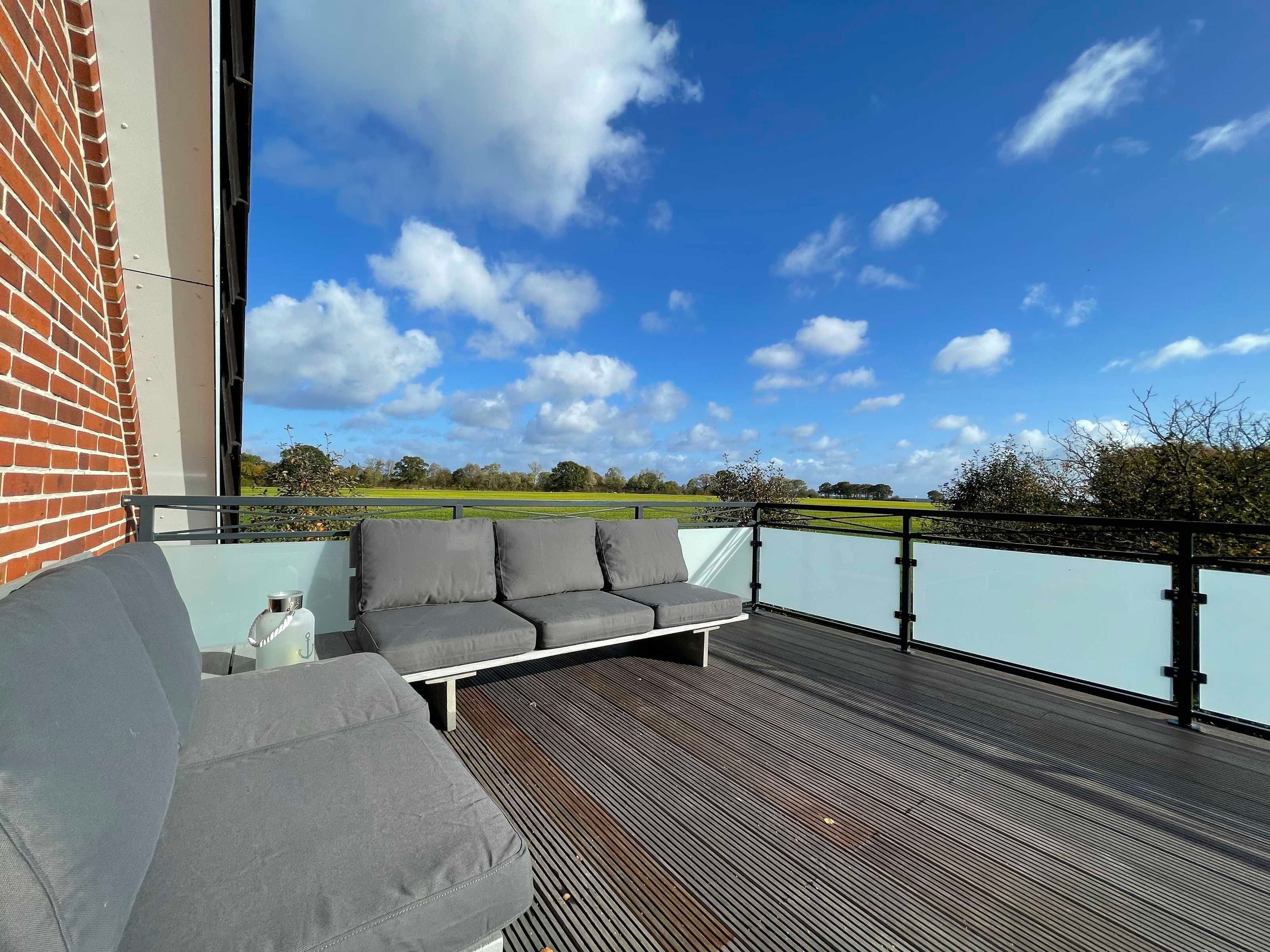Een balkon met een grijze bank en een kleine tafel, met uitzicht op een groen veld en een helderblauwe lucht.