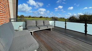 A balcony with a grey sofa and a small table, overlooking a green field and a clear blue sky. - Vacation home Days by the sea, 3 bedrooms, 3 bathrooms, with roof terrace - Holiday home with 3 bedrooms (Kellenhusen (Ostsee))