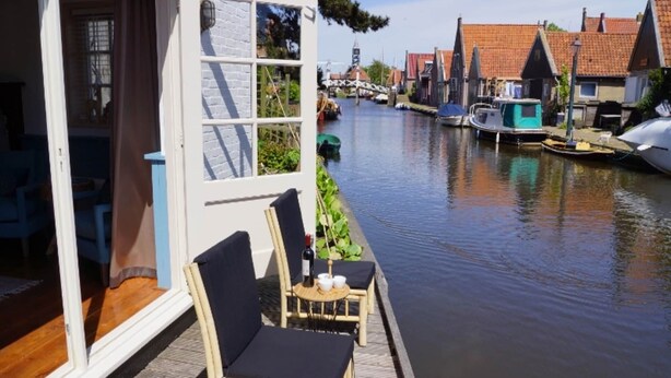 A wooden deck with two wicker chairs and a small table, overlooking a canal with boats and traditional buildings in the background. - Holiday Home in Hindeloopen by Lake (Hindeloopen)