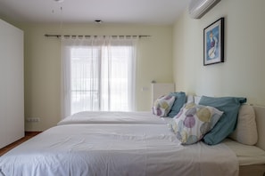 A room with two beds, a window with sheer curtains, a wall-mounted air conditioner, and a framed picture.