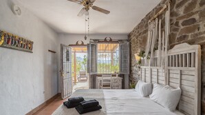 A bedroom with a bed, a ceiling fan, a stone wall, and a view through open doors. - Holiday Home 'Finca Arrijana' with Mountain View, Wi-Fi and Air Conditioning (Sayalonga)