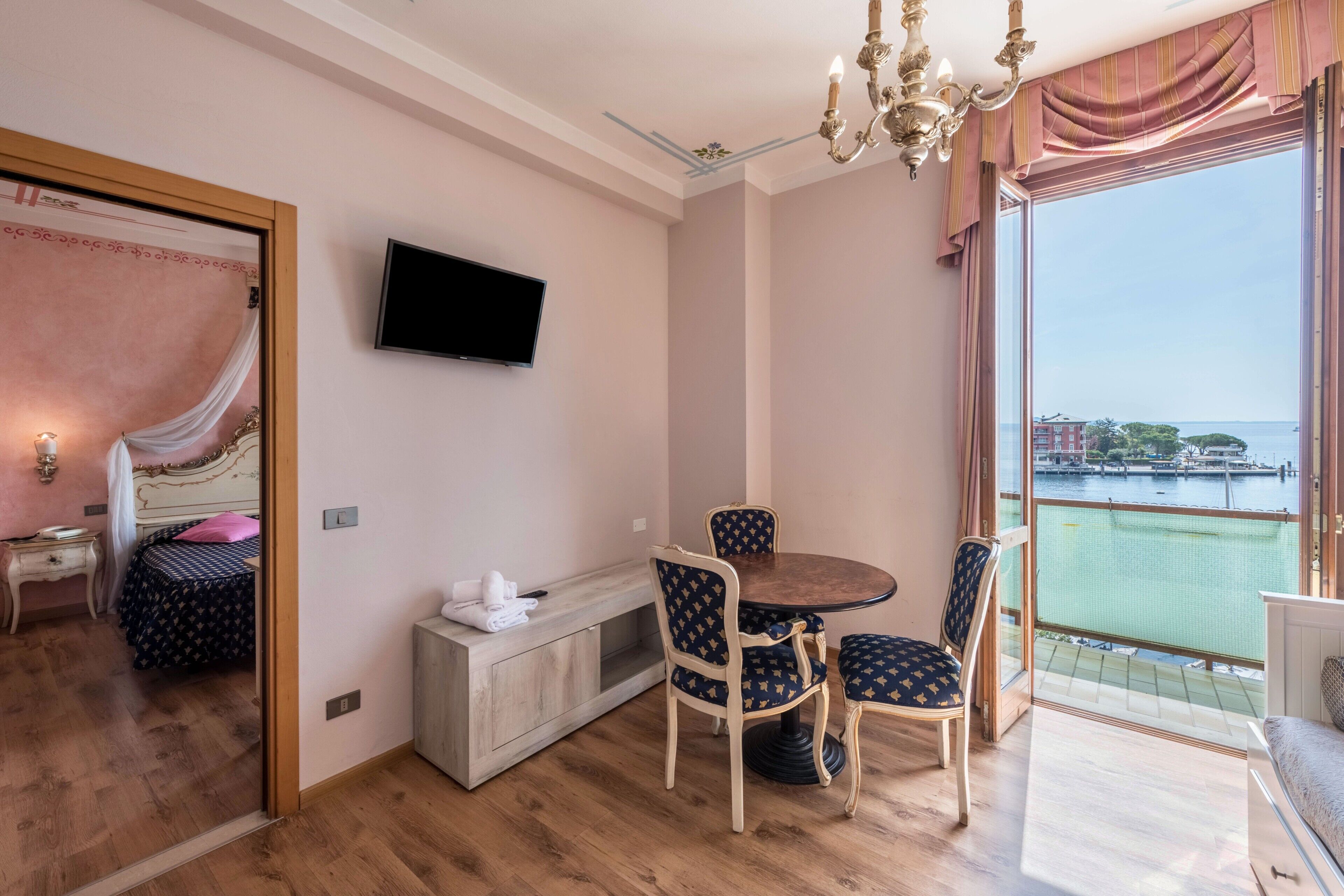 Une chambre avec un lit, une télévision, une table à manger avec des chaises, un balcon donnant sur l’eau et un lustre.