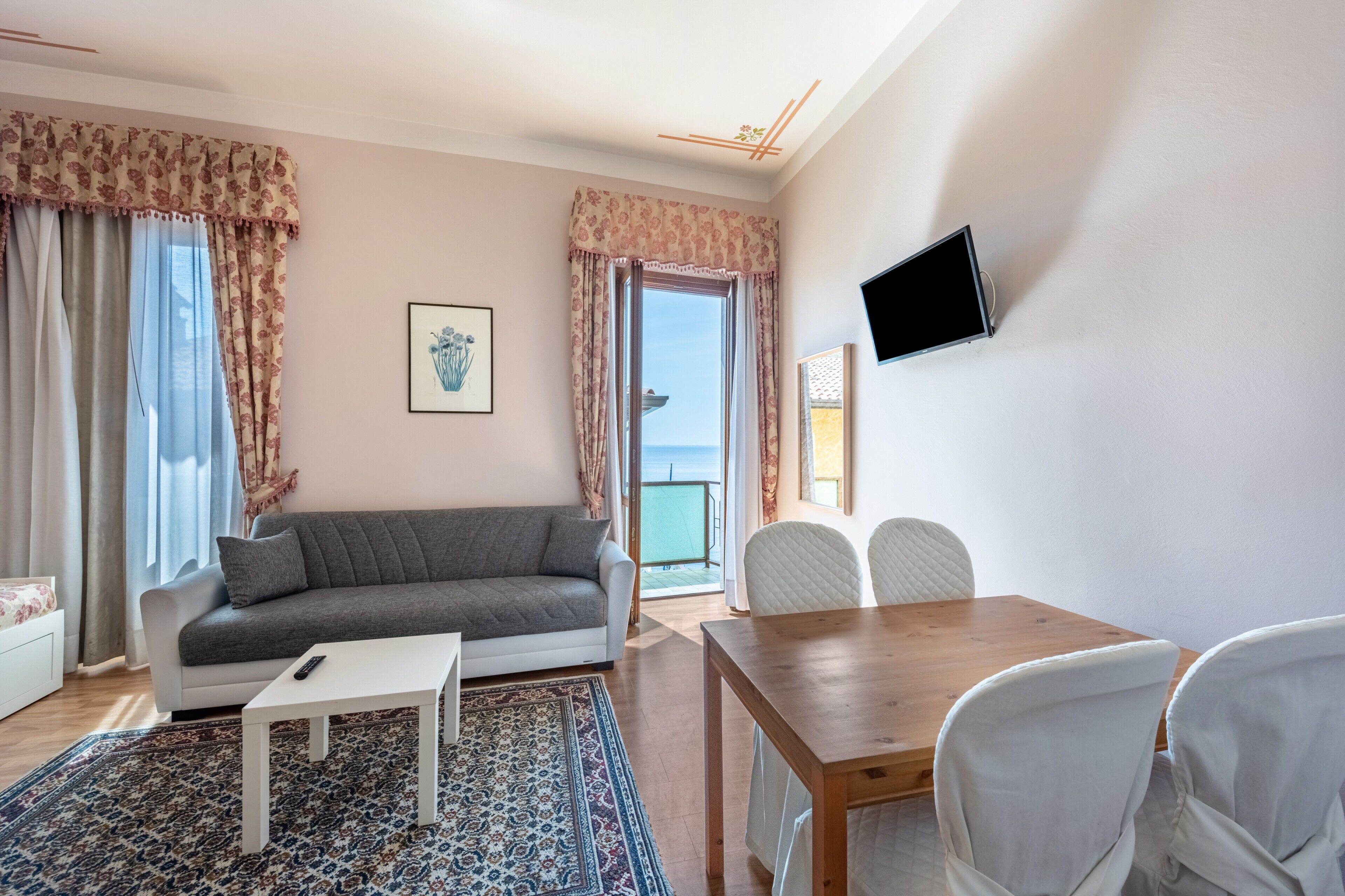 Ein Wohnzimmer mit Sofa, Couchtisch, Esstisch mit Stühlen, einem an der Wand befestigten Fernseher und Meerblick durch das Fenster.