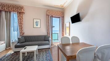 Ein Wohnzimmer mit Sofa, Couchtisch, Esstisch mit Stühlen, einem an der Wand befestigten Fernseher und Meerblick durch das Fenster.