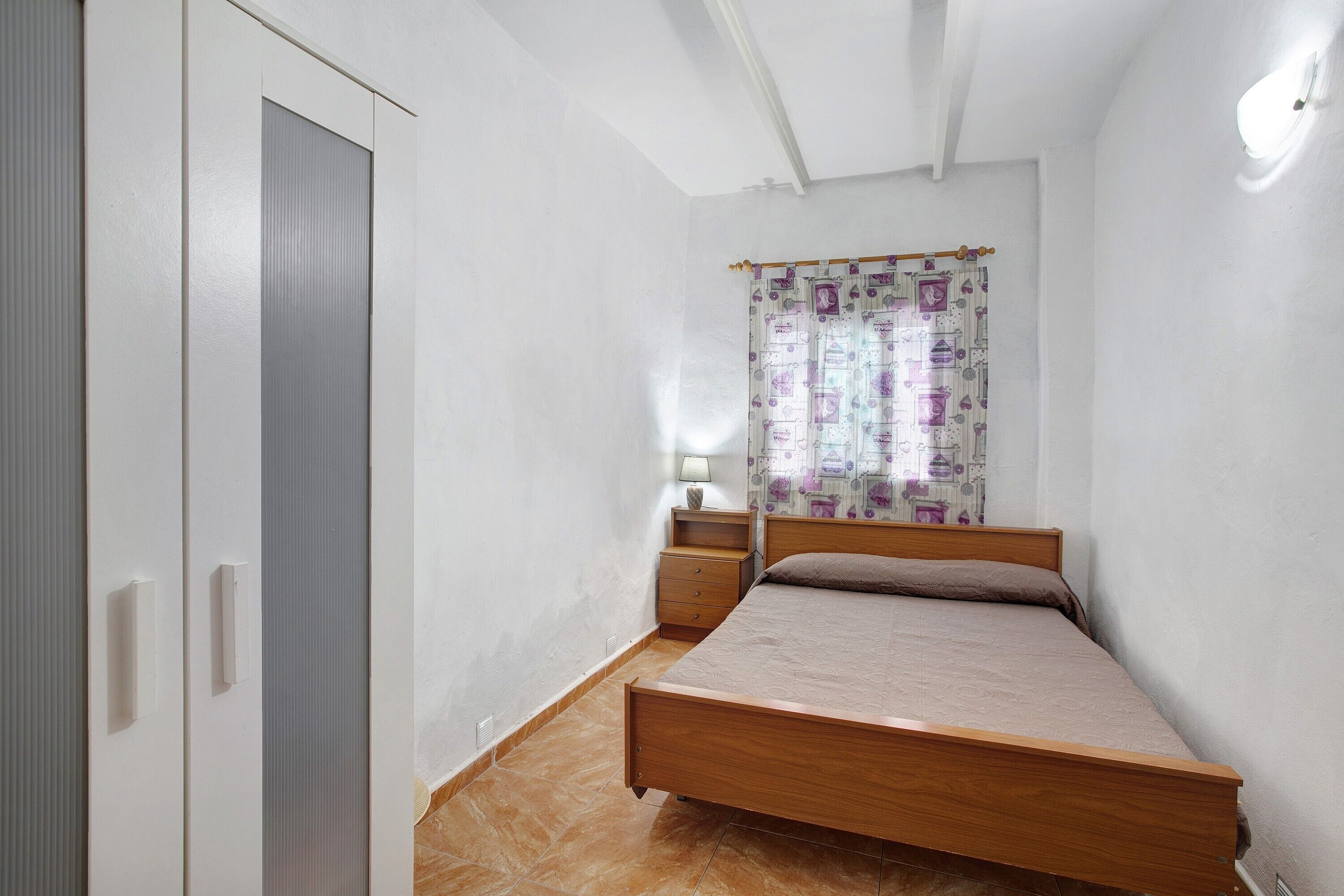 A bedroom with a bed, a wardrobe, a nightstand, a lamp, and a curtain.