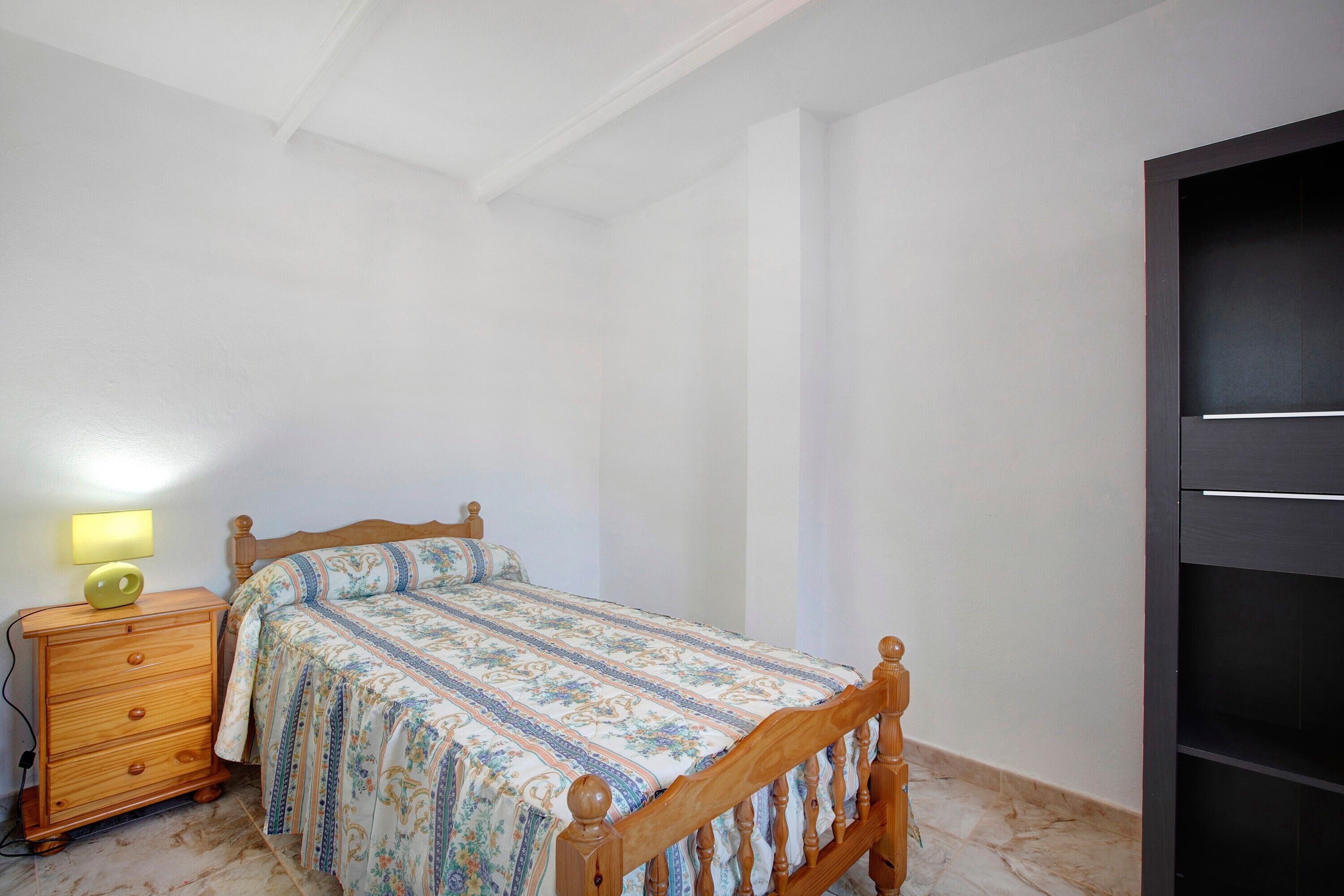 A bedroom with a bed, a wooden dresser, a nightstand with a lamp, and a dark wardrobe.