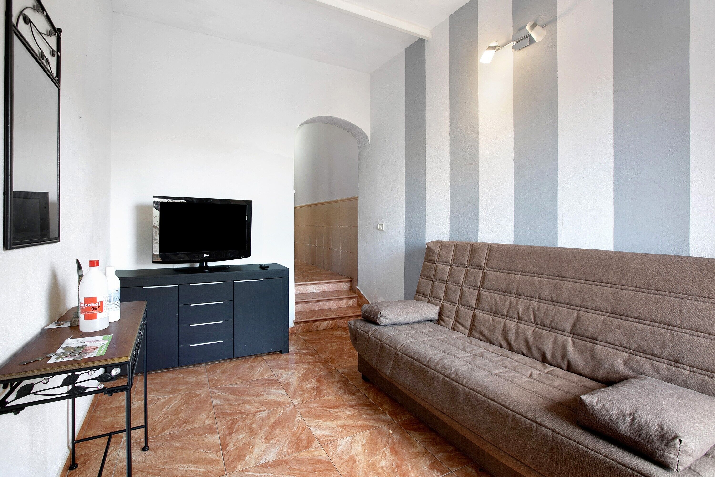 A living room with a sofa, a TV on a stand, a table with bottles, and a staircase in the background.