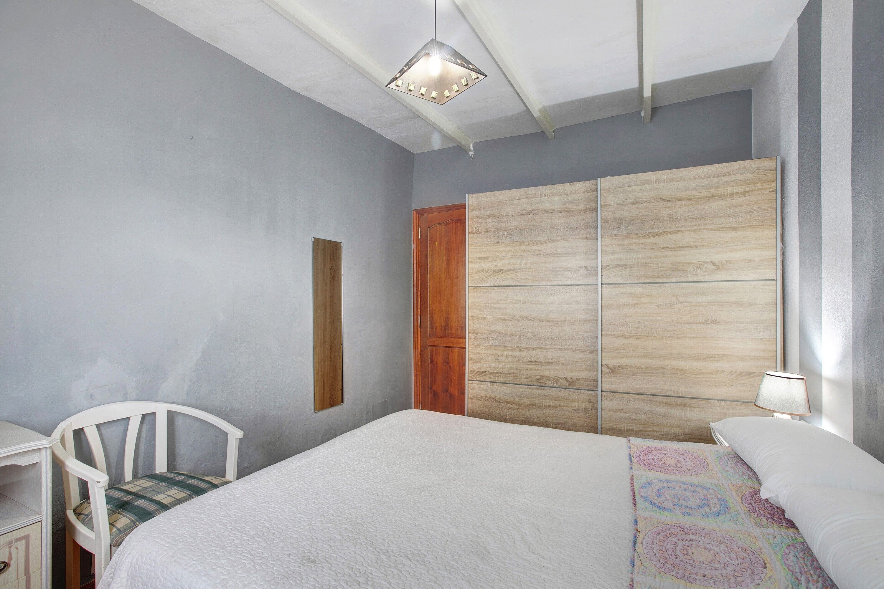 A bedroom with a bed, a chair, a wooden wardrobe, a lamp, and a ceiling light fixture.