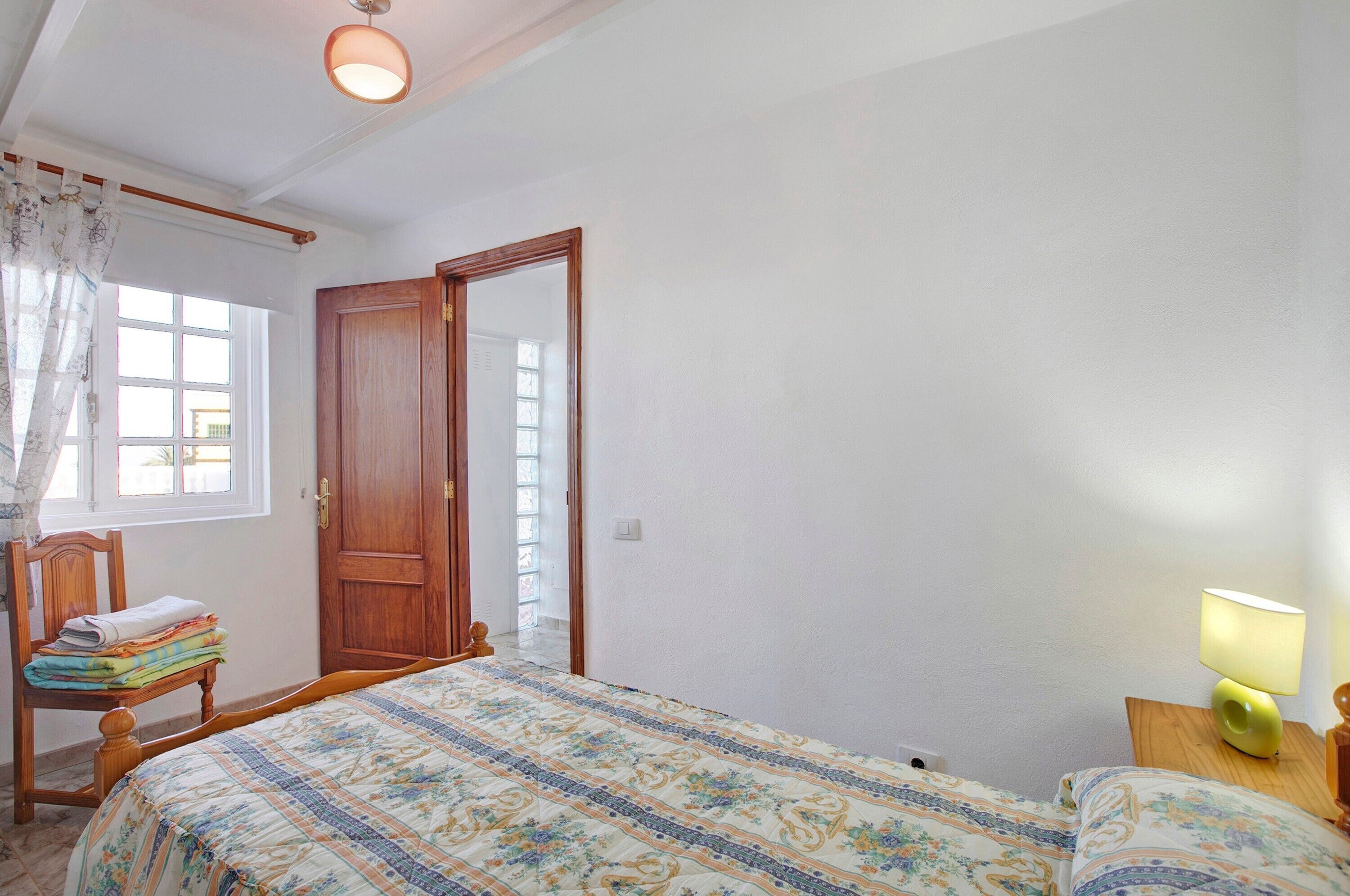 Um quarto com cama, uma cadeira com toalhas dobradas, uma porta de madeira e uma mesa de cabeceira com abajur.