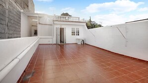 Una terraza en la azotea con suelo de baldosas, un edificio blanco con balcón y cielo despejado.