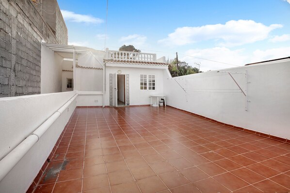 A rooftop terrace with a tiled floor, a white building with a balcony, and a clear sky.