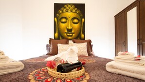 A bed with a patterned bedspread, a Buddha painting on the wall, a wooden wardrobe, and a basket with towels and a bottle. - Vacation Home "Casa Alexis" with Wi-Fi, Terrace & Garden (La lajita)