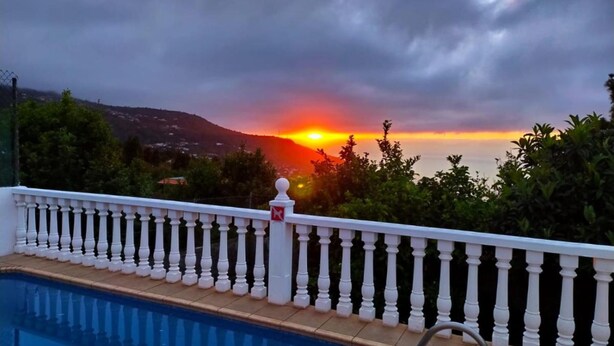 Una piscina con balaustrada blanca, vista al atardecer sobre el mar y un paisaje montañoso al fondo.