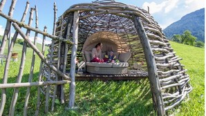 Una estructura de madera rústica con techo tejido, un espacio de descanso acolchado y una pequeña mesa.