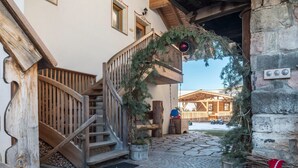 A wooden staircase leading to a balcony with a wooden railing, a stone wall, and a wooden building in the background.