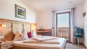 A wooden bed with floral bedding, a nightstand, a desk, a chair, a balcony with a railing, and a window with curtains.
