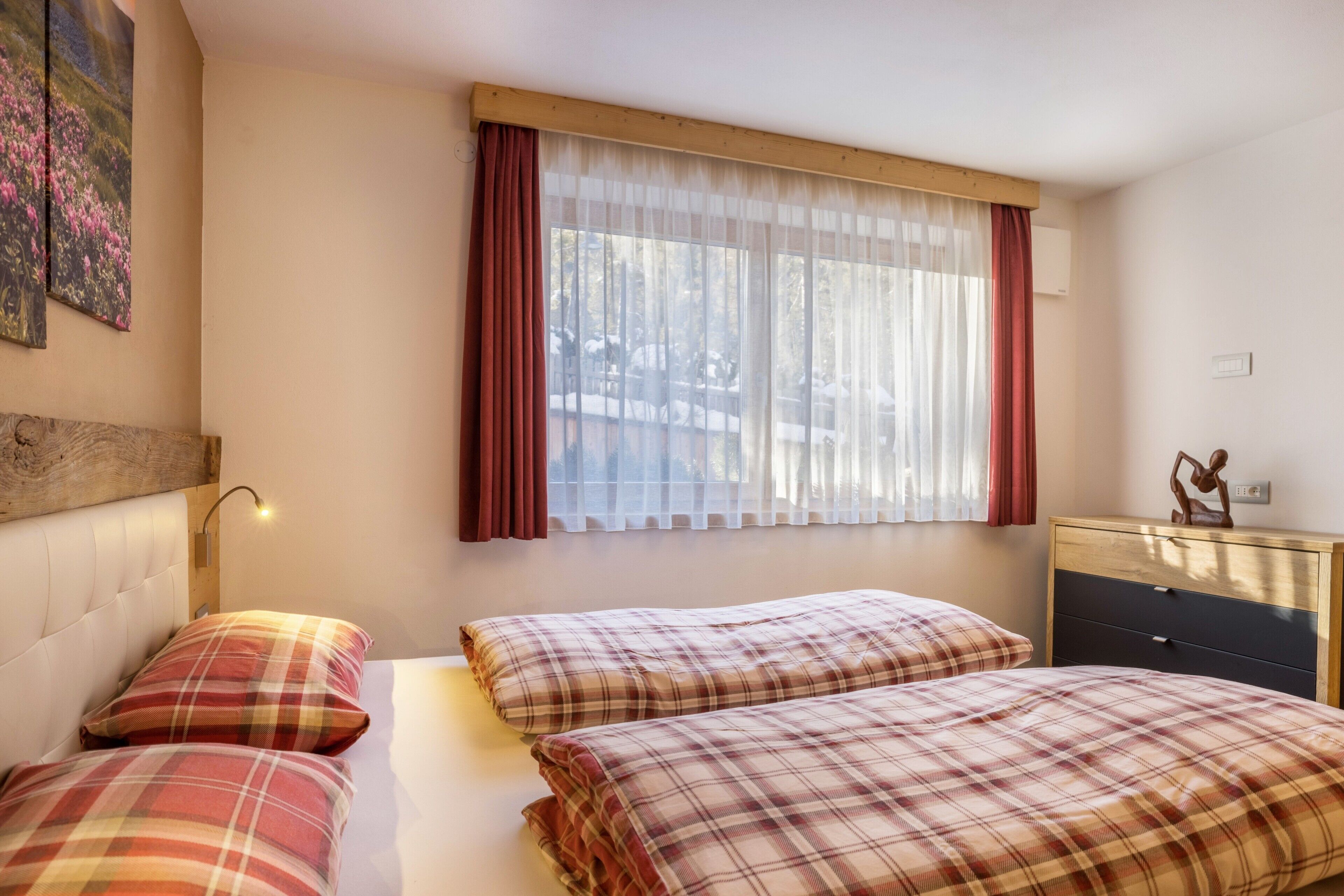 Une chambre avec deux lits, une fenêtre avec des rideaux légers, une tête de lit en bois, une table de chevet avec une lampe et une petite commode.