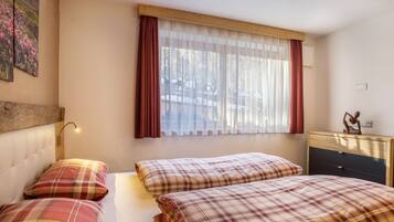 Une chambre avec deux lits, une fenêtre avec des rideaux légers, une tête de lit en bois, une table de chevet avec une lampe et une petite commode.