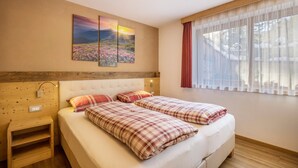 A bedroom with two beds, a wooden headboard, a nightstand, a wall-mounted lamp, and a window with sheer curtains. - Apartment 'Ciasa Rungg App Pares' with Mountain View, Garden & Wi-Fi (St.Vigil)