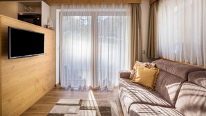 A living room with a flat-screen TV, wooden paneling, a sofa with cushions, a patterned rug, and a sliding glass door with sheer curtains. - Apartment 'Ciasa Rungg App Pares' with Mountain View, Garden & Wi-Fi (St.Vigil)
