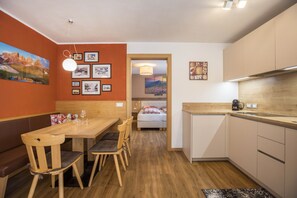 A compact hotel room with a wooden dining table, chairs, a bed, and a kitchenette.