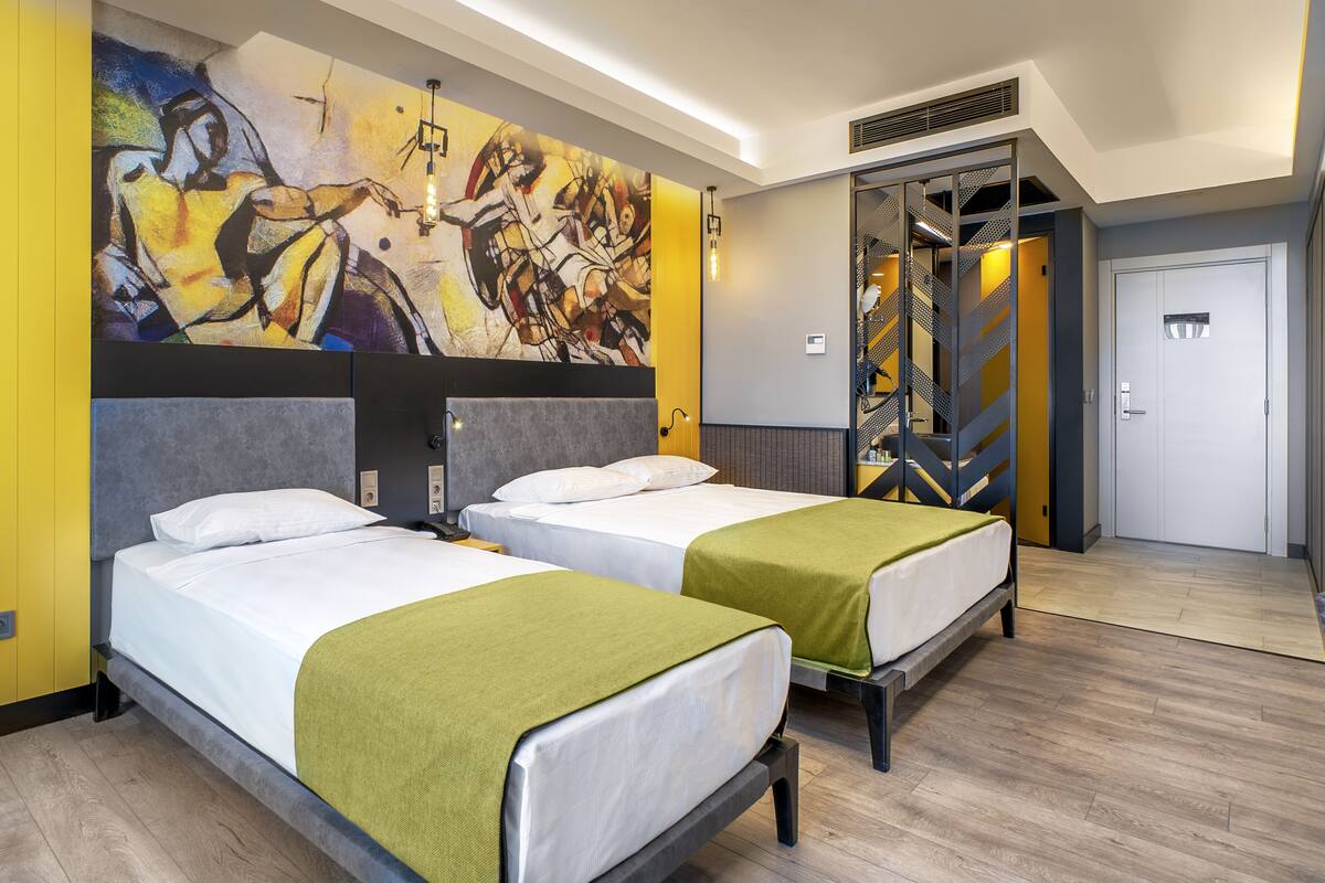 Hotellihuone, jossa on kaksi sänkyä, suuri abstrakti maalaus seinällä ja portaikko, joka johtaa toiseen huoneeseen.