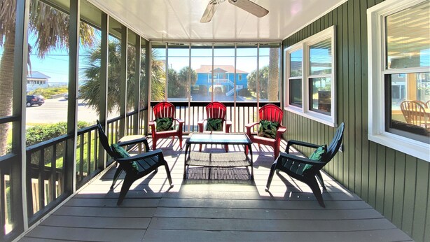 A covered porch with a ceiling fan, red chairs with green cushions, a table, and large windows offering a view of the street and neighboring houses.