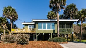 A two-story house with a balcony, surrounded by pine straw and palm trees.