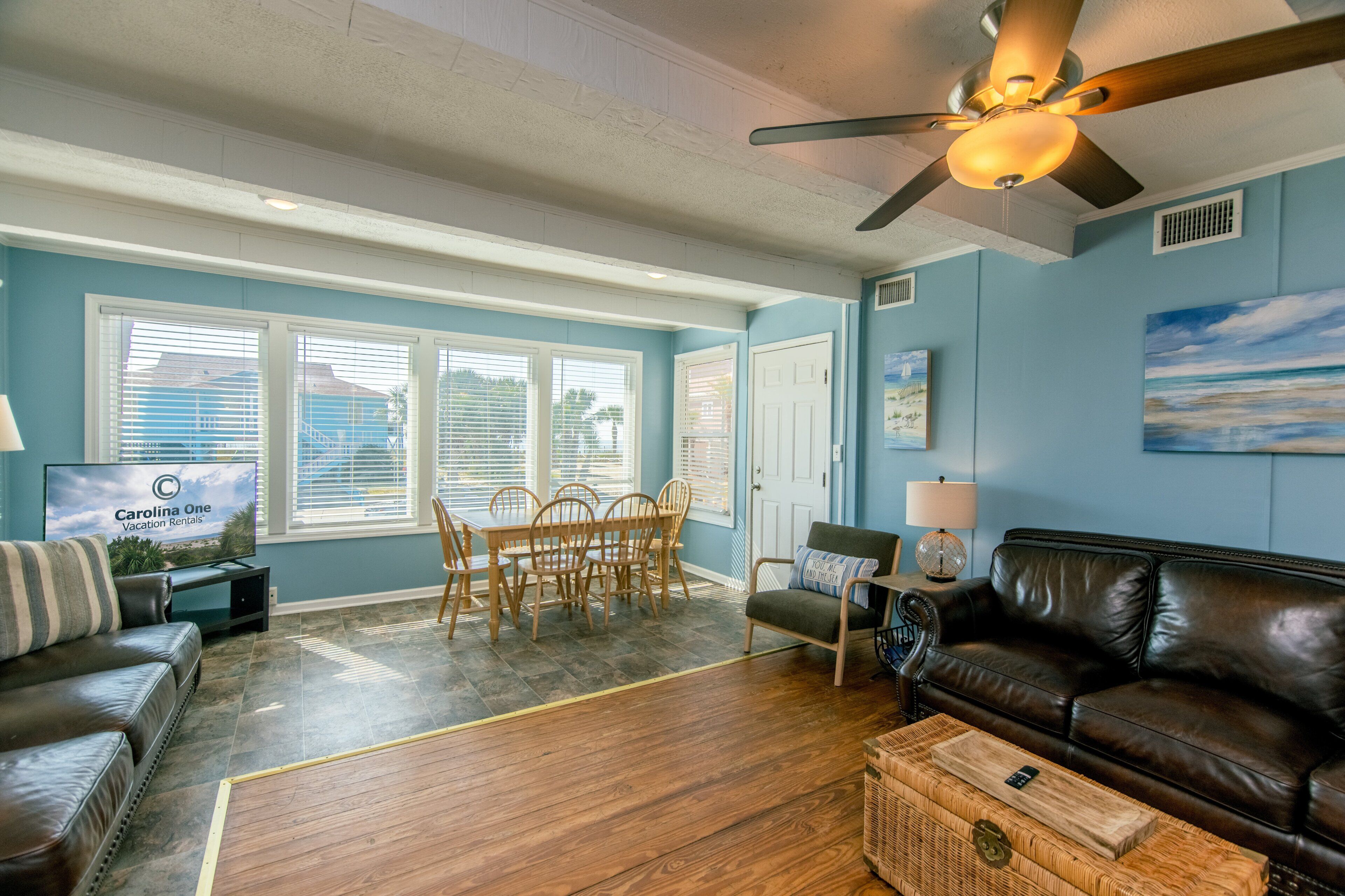 A living room with a ceiling fan, a brown leather couch, a dining table with chairs, and a television displaying 'Carolina One Vacation Rentals'.